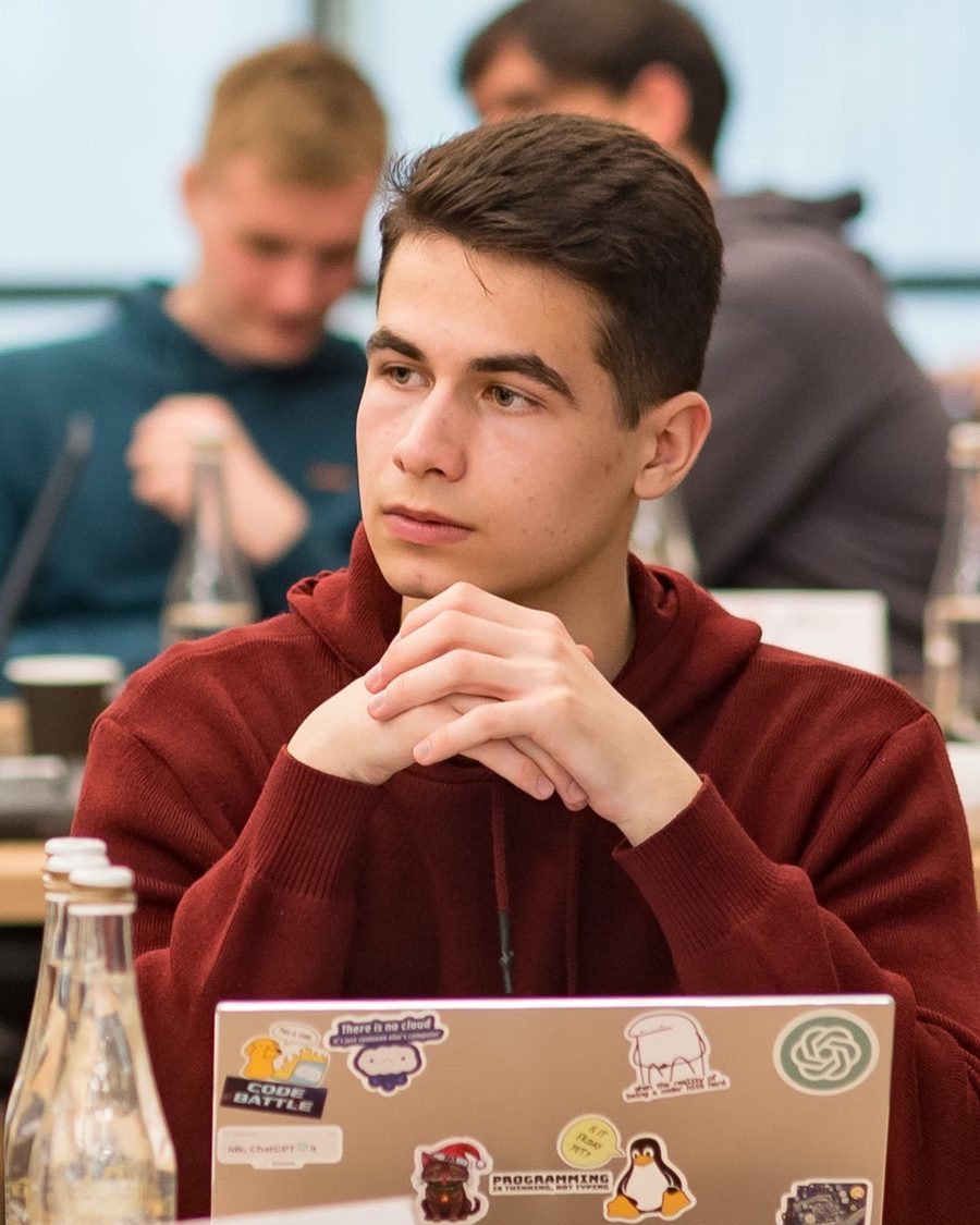 Maxim Isacescu in a red hoodie, seated at a table with a laptop covered in programming stickers